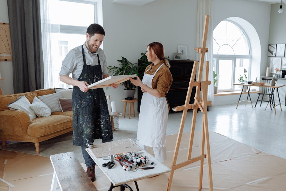 Two artists in aprons discussing art in a sunlit studio with canvases and easels.